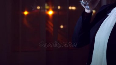 İş takım elbiseli genç bir kadın geceleri bir ev pencerenin önünde duran kırmızı şarap içiyor. Pencere panorama, gece ışık şehir görüntüsü. 4k