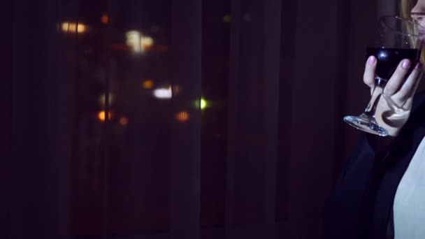 Μια νεαρή γυναίκα σε ένα επαγγελματικό κοστούμι πόσιμο κόκκινο κρασί, στέκεται στο παράθυρο του ένα σπίτι το βράδυ. Παράθυρο Πανόραμα, θέα στην πόλη, φώτα τη νύχτα. 4k — Αρχείο Βίντεο