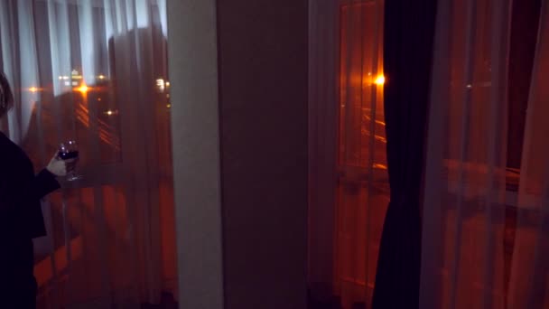 Una joven con un traje de negocios está bebiendo vino tinto, de pie en la ventana de una casa por la noche. Ventana panorámica, vista de la ciudad, luces nocturnas. 4k — Vídeos de Stock