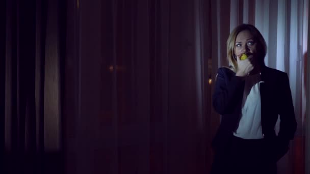 Mujer en un traje de negocios comiendo una manzana parada junto a la ventana panorámica de la noche. vista de la ciudad, luces nocturnas. 4k — Vídeo de stock