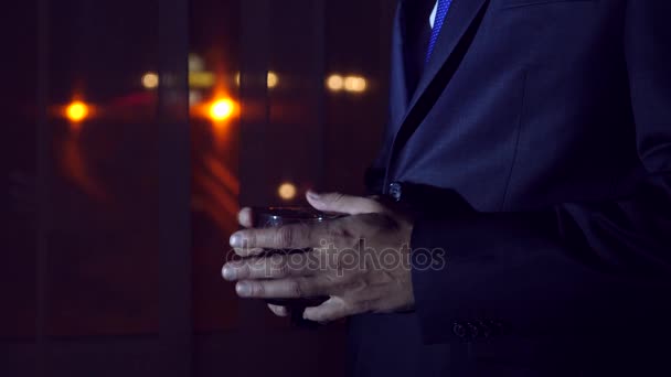 A young man in a business suit is drinking alcohol, standing at the window of a house at night. Window panorama, view of the city, night lights. 4k — Stock Video