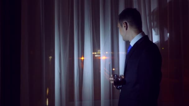 A young man in a business suit is drinking alcohol, standing at the window of a house at night. Window panorama, view of the city, night lights. 4k — Stock Video
