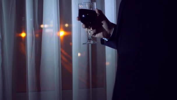 Ein junger Mann im Businessanzug trinkt Alkohol und steht nachts am Fenster eines Hauses. Fensterpanorama, Blick auf die Stadt, Nachtbeleuchtung. 4k — Stockvideo