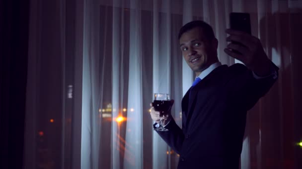 Молодий чоловік у діловому костюмі п'є алкоголь і робить селфі на своєму смартфоні, стоїть біля вікна будинку вночі. Панорама вікон, вид на місто, нічні вогні. 4k — стокове відео