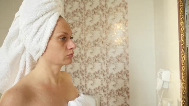 Женщина наносит увлажняющий крем для кожи на лицо, глядя в зеркало в ванной комнате. 4k, slow motion — стоковое видео