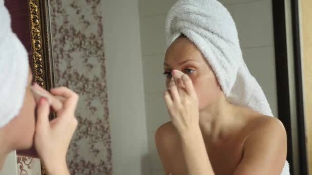 Женщина наносит увлажняющий крем для кожи на лицо, глядя в зеркало в ванной комнате. 4k, slow motion — стоковое видео