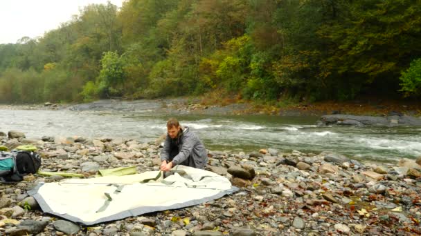 4k. der Typ stellt ein Touristenzelt am Ufer eines Gebirgsflusses auf. — Stockvideo