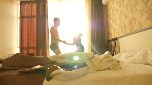 A cheerful couple dancing and jumping for joy in front of the panoramic window of the bedroom. sun glare from the window. 4k. Slow motion — Stock Video