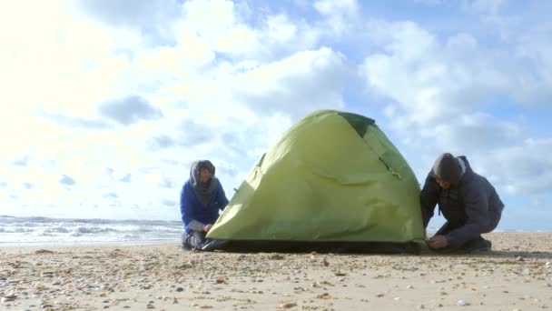 Tenda de acampamento na praia junto ao mar. 4K, câmara lenta. um homem e uma mulher montam uma barraca no tempo ventoso em uma praia arenosa — Vídeo de Stock