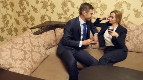 Двое бизнесменов, мужчина и женщина в деловых костюмах пьют вино в люксах, сидят на диване. 4k — стоковое видео