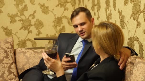 Пара бізнесменів, чоловік і жінка в ділових костюмах п'ють вино в сюїтах, сидячи на дивані. 4k — стокове відео