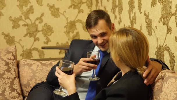 Двое бизнесменов, мужчина и женщина в деловых костюмах пьют вино в люксах, сидят на диване. 4k — стоковое видео
