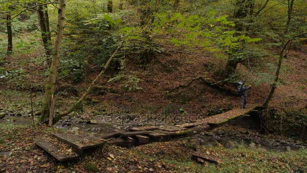 Un turista masculino está caminando a lo largo de un cruce hecho a sí mismo sobre un río de montaña en un bosque más. 4k, cámara lenta — Vídeo de stock