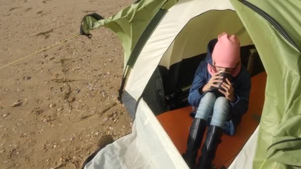 La chica se sienta en una tienda de campaña y bebe una bebida caliente de una taza termo. Tienda de campaña en la playa junto al mar. 4k, cámara lenta . — Vídeos de Stock