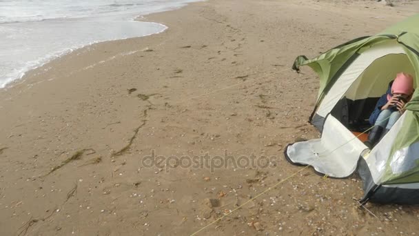 Sitzt das Mädchen in einem Zelt und trinkt ein Heißgetränk aus einem Thermobecher. Zeltlager am Strand am Meer. 4k, Zeitlupe. — Stockvideo