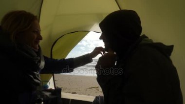 bir erkek ve bir kadın bir sıcak kupa ve öpücük, bir çadır Denizi üzerinde bir bakış zemin karşı sıcak çay iç. Denizin kenarında plajda kamp. 4k, ağır çekim.