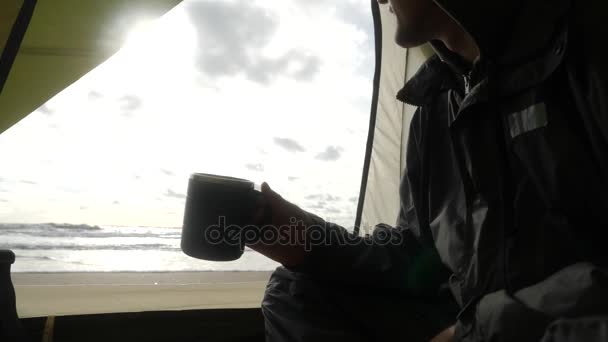 El hombre está bebiendo té caliente de una taza termo contra el telón de fondo de la vista desde la tienda en el mar. Acampar en la playa junto al mar. 4k, cámara lenta . — Vídeos de Stock