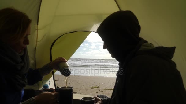 Vor dem Hintergrund des Blicks aus dem Zelt auf das Meer trinken der Mann und die Frau heißen Tee aus einem Thermobecher. Zelten am Strand am Meer. 4k, Zeitlupe. — Stockvideo