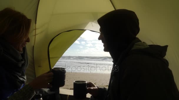 Um homem e uma mulher bebem chá quente de uma caneca quente e beijam, no pano de fundo de um vislumbre de uma tenda no mar. Acampar na praia junto ao mar. 4k, câmera lenta . — Vídeo de Stock