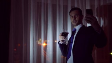 Genç bir adam bir iş takım elbise içecekler alkol ve onun akıllı yapar selfie geceleri evi pencerenin önünde duruyor. Pencere panorama, gece ışık şehir görüntüsü. 4k