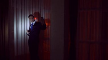 Genç bir adam bir iş takım elbise içecekler alkol ve onun akıllı yapar selfie geceleri evi pencerenin önünde duruyor. Pencere panorama, gece ışık şehir görüntüsü. 4k