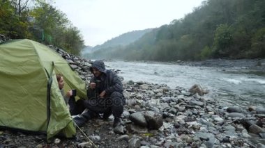 bir turist çadır 4 k. görünümünden bir dağ nehir üzerinde. Kız adamla çay termosu şişe içine dökülen ve dağ nehir görünüyor.