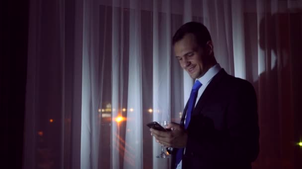 Ein junger Mann im Businessanzug trinkt Alkohol und macht ein Selfie mit seinem Smartphone, steht nachts am Fenster des Hauses. Fensterpanorama, Blick auf die Stadt, Nachtbeleuchtung. 4k — Stockvideo