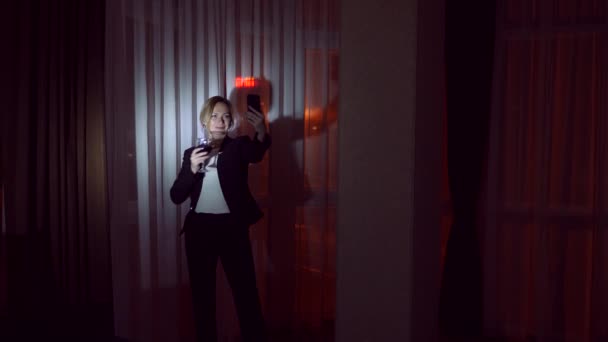 Una mujer en un traje de negocios bebe alcohol, y hace selfie en su teléfono inteligente, se para en la ventana de la casa por la noche. Ventana panorámica, vista de la ciudad, luces nocturnas. 4k — Vídeos de Stock