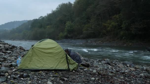 4K. um cara perto de uma tenda turística na margem de um rio de montanha . — Vídeo de Stock