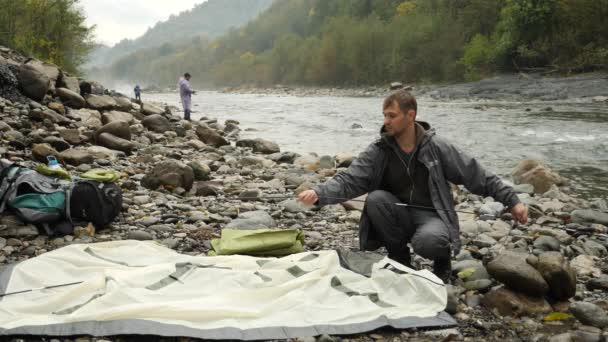 4k. el tipo pone una tienda de campaña turística en la orilla de un río de montaña . — Vídeos de Stock