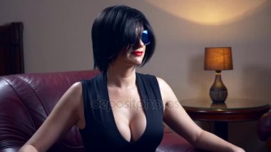 Sıcak esmer kadın güneş gözlüğü yuvarlak, bir sandalyeye oturur ve kameraya benziyor. 4k, ağır çekim