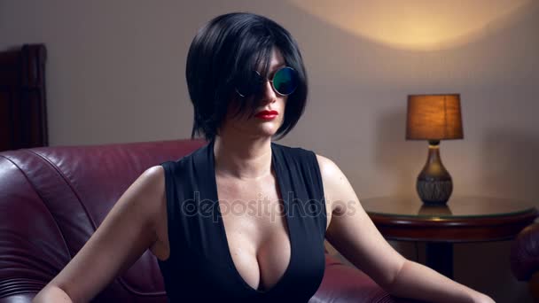 Hot brunette woman in round sunglasses, sits in a chair and looks at the camera. 4k, slow motion — Stock Video