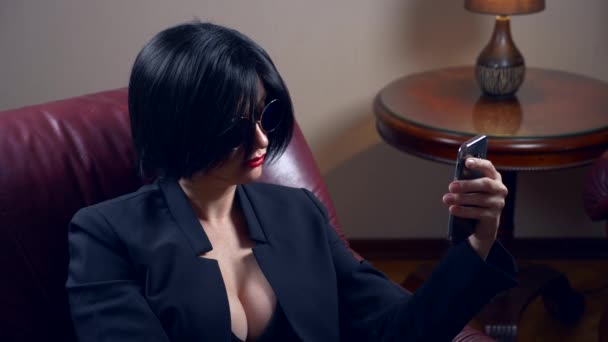 Heiße Brünette mit runder Sonnenbrille, sitzt in einem Sessel und schreibt eine Nachricht auf ihr Smartphone. 4k, Zeitlupe — Stockvideo