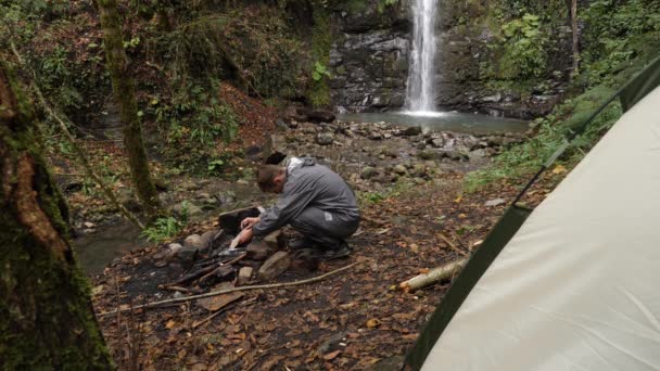 4k, slowmotion. manliga turist bränder en brasa för en grill i skogen mot bakgrund av ett vattenfall. — Stockvideo