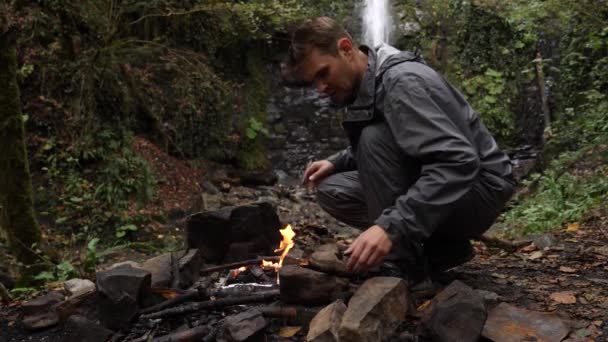 4k, slow motion. a male tourist fires a bonfire for a grill in the forest against the backdrop of a waterfall. — Stock Video