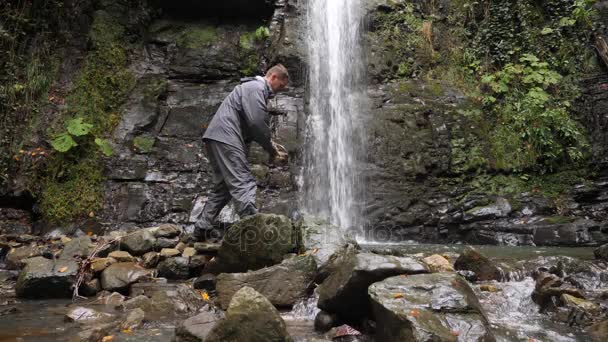 4k, slowmotion. en turist man är att plocka upp vatten från en sjö vid foten av ett berg bredvid ett vattenfall. — Stockvideo