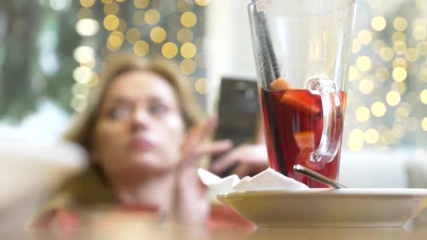 Красива жінка відпочиває в кафе, п'є гаряче глінтвейн і заглядає в свій смартфон. 4k, розмивання тла . — стокове відео