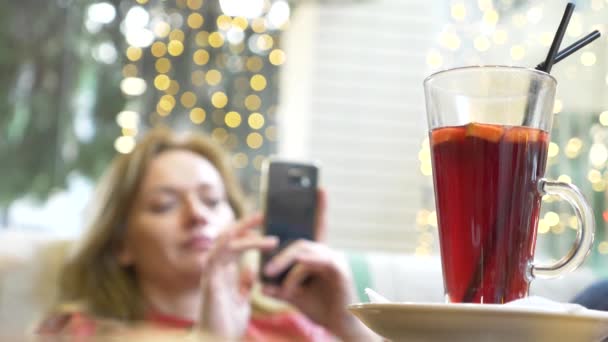 Hermosa mujer está descansando en un café, beber vino caliente caliente con especias y mirando en su teléfono inteligente. 4k, desenfoque de fondo . — Vídeo de stock