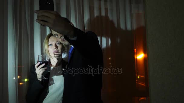 Uma jovem mulher em um terno de negócios e um homem fazendo uma gravação de vídeo em seu smartphone para redes sociais, de pé à janela de uma casa à noite. Panorama da janela, vista da cidade, luzes noturnas — Vídeo de Stock