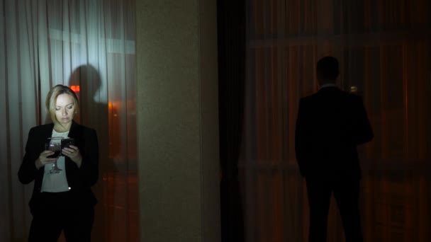 Молода жінка в діловому костюмі і чоловік розмовляє, стоячи біля вікна будинку вночі. Панорама вікон, вид на місто, нічні вогні. 4k — стокове відео