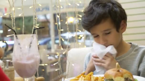 El chico está comiendo una hamburguesa en un café. 4k. una hamburguesa con una chuleta . — Vídeos de Stock