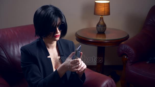 Горячая брюнетка в круглых солнцезащитных очках, сидит в кресле и пишет сообщение на своем смартфоне. 4k, slow motion — стоковое видео