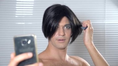 Travesti makyaj. genç bir adam kendine bir transseksüel mencap hazırlar. 4k, ağır çekim. o peruk üzerinde koyar ve sosyal ağ kendi Smartphone'da koyar