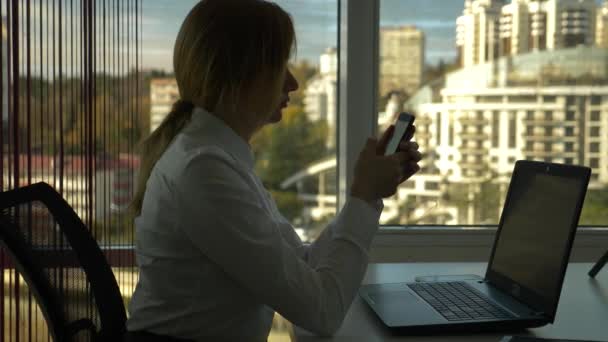 Mulher de negócios em um escritório moderno com uma janela panorâmica, dia ensolarado, laptop, tablet, use um telefone celular em uma situação. 4k, câmera lenta — Vídeo de Stock