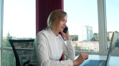 iş kadını bir panoramik pencere, güneşli bir gün, dizüstü bilgisayar, tablet, modern bir ofiste bir durumda bir cep telefonu kullanın. 4k, ağır çekim