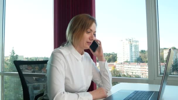 商务妇女在一个现代化的办公室里有一个全景窗口, 阳光明媚的日子, 笔记本电脑, 平板电脑, 在一个情况下使用手机。4k、慢动作 — 图库视频影像