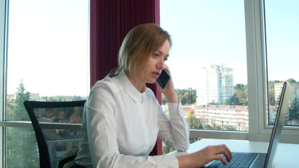 Mulher de negócios em um escritório moderno com uma janela panorâmica, dia ensolarado, laptop, tablet, use um telefone celular em uma situação. 4k, câmera lenta — Vídeo de Stock