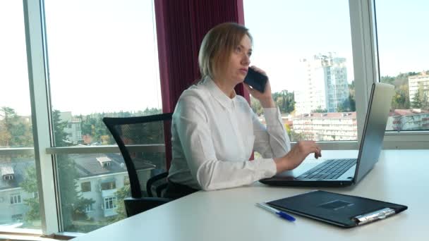 Femme d'affaires dans un bureau moderne avec une fenêtre panoramique, journée ensoleillée, ordinateur portable, tablette, utiliser un téléphone mobile dans une situation. 4k, au ralenti — Video