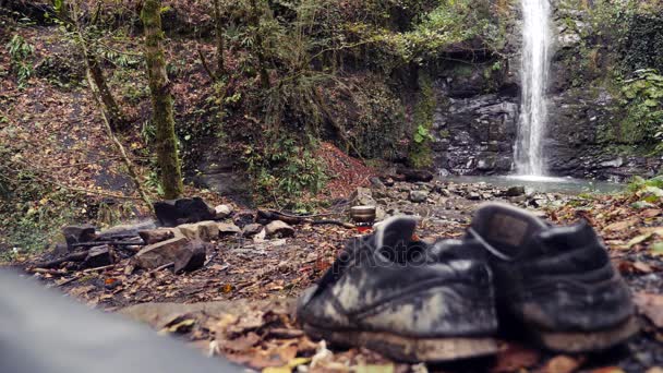 4k, lassú mozgás. A háttérben egy vízesés, a hegy lábánál a piszkos turista cipő — Stock videók