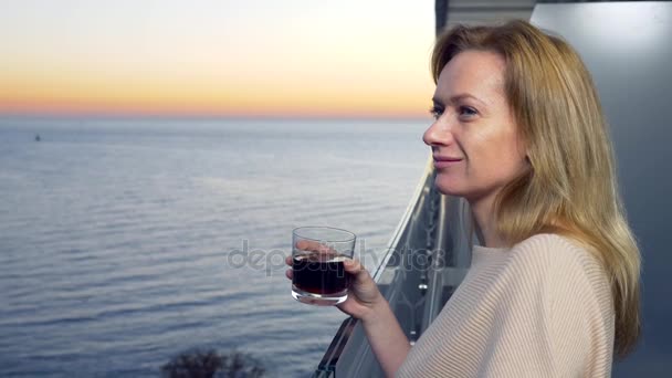 여자는 바다 전망의 리조트 발코니에 서 있다. 일몰을 보고 저녁에 칵테일을 마신다. 4 k — 비디오